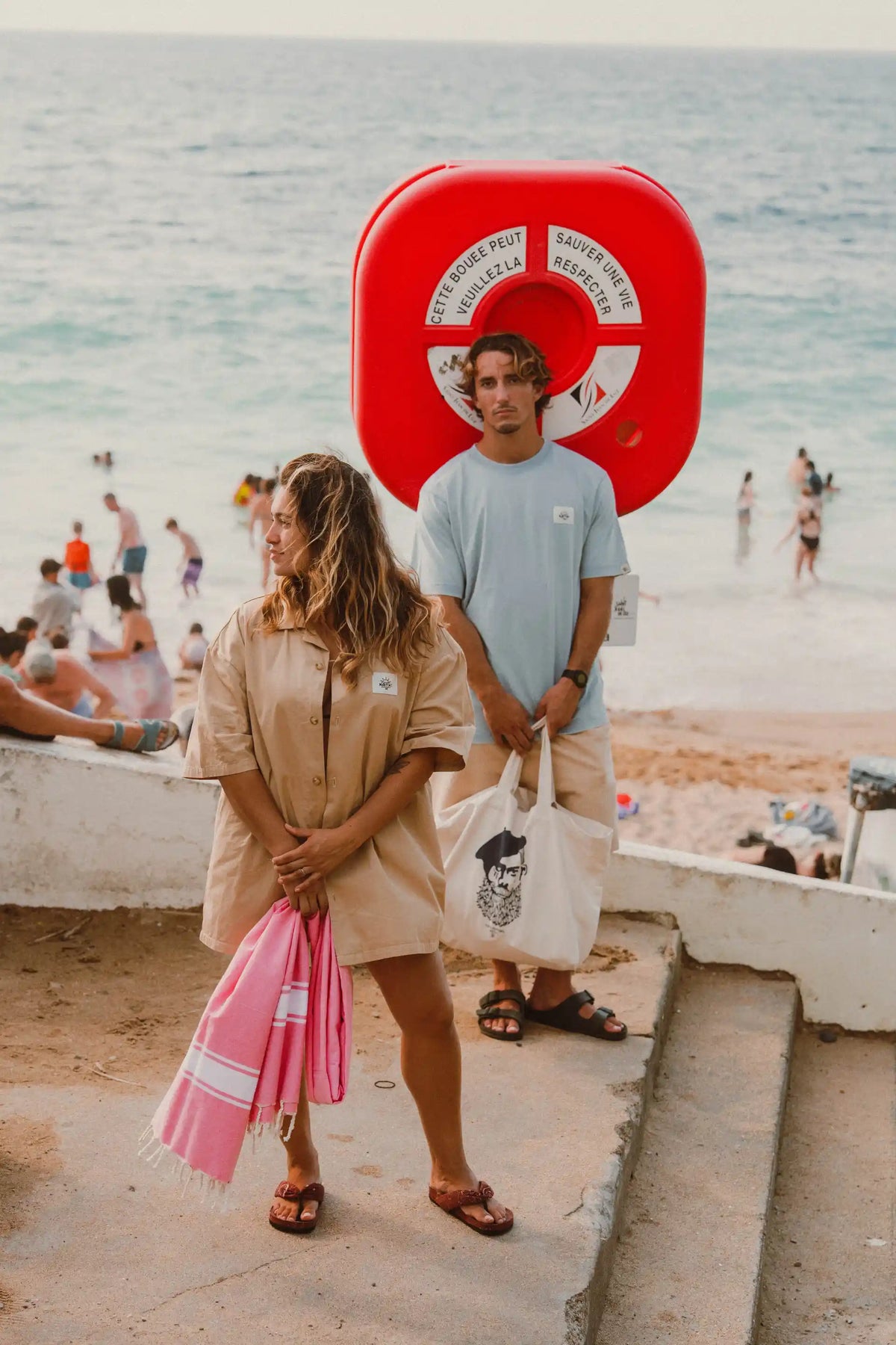 Couple en tenues Katxi Klothing sur la plage devant une bouée de sauvetage