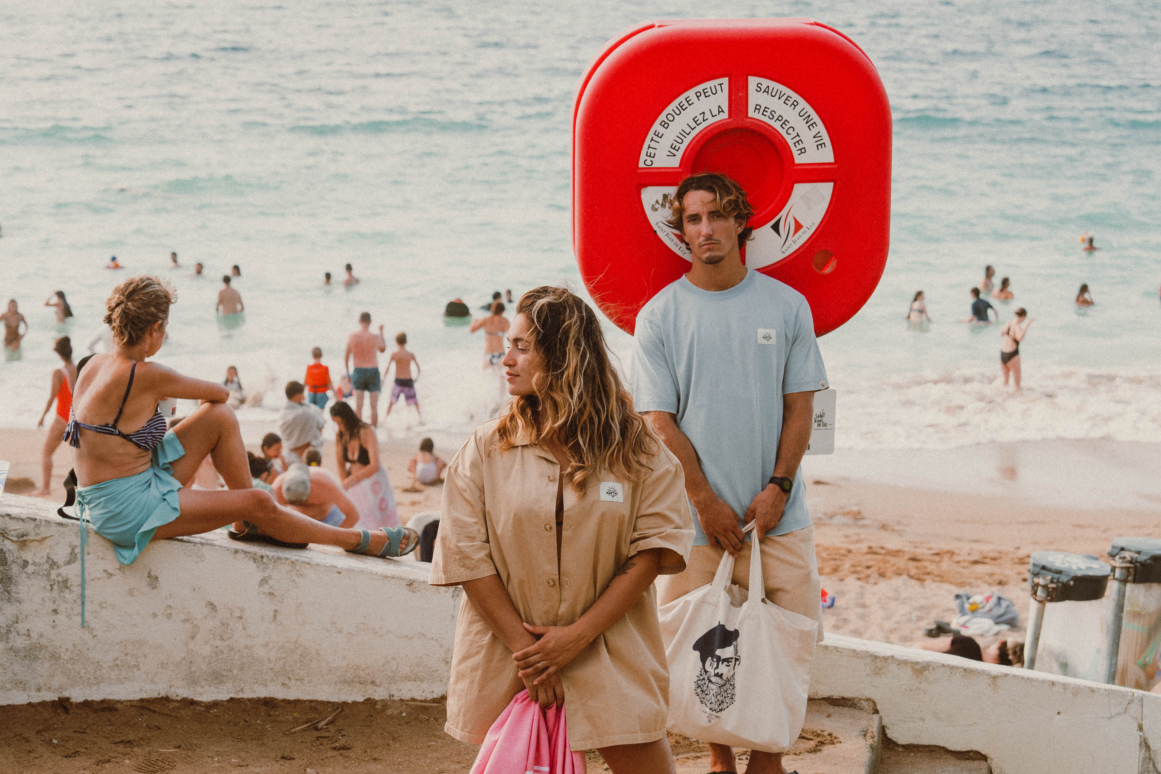 Couple en tenues Katxi Klothing sur la plage devant une bouée de sauvetage
